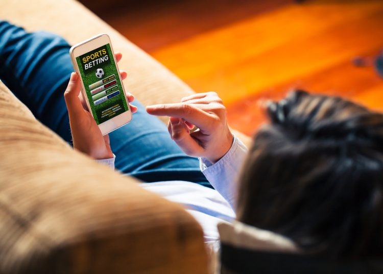 Woman holding a mobile phone to visit a sports betting website while lies down at home.